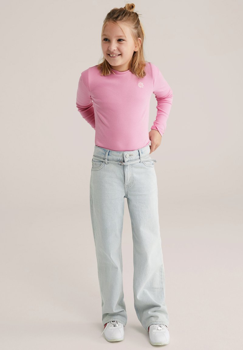 Cămașă roz cu mâneci lungi, cu dungi, asortată cu blugi largi, de culoare albastru deschis. Figura stă cu mâinile pe șold, purtând adidași albi.