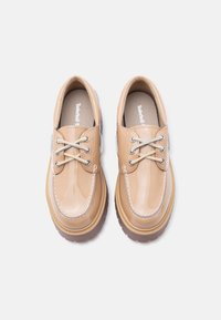 Μπεζ παπούτσια από δερμάτινο λουστρίνι με στρογγυλεμένη μύτη, αντίθετη λευκή ραφή και ελαφριά καφέ καουτσούκ σόλα· διαθέτουν παραδοσιακό δέσιμο με κορδόνια.