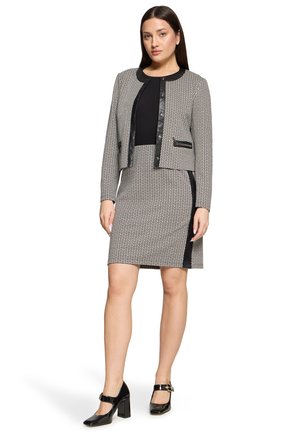 Femme portant un haut noir avec une veste à motif et une jupe assortie jusqu'aux genoux, accompagnée de chaussures Mary Jane noires à talons hauts.