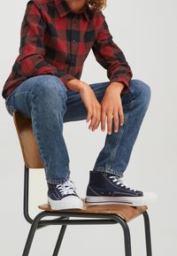 Röd och svart rutad skjorta, blå jeans och marinblå högskor med vita sulor. Person sitter på en trästol.