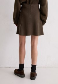 Persoon draagt een bruine wollen rok, bijpassende bruine trui, zwarte sokken en bruine leren schoenen, staand tegen een witte textuurmuur.