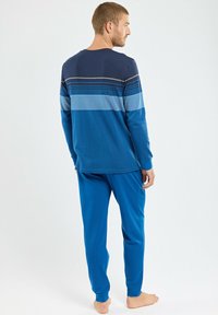 Homme debout pieds nus portant un haut à manches longues rayé bleu et un pantalon bleu assorti, de profil contre un fond uni.