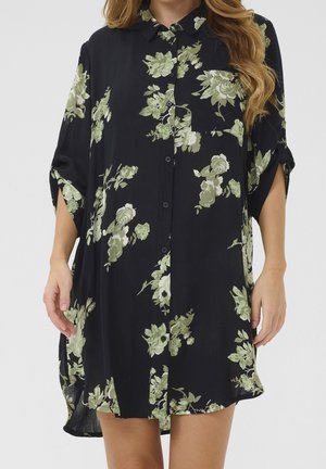Femme portant une robe noire à boutons avec manches retroussées et imprimé floral vert, debout les mains détendues le long du corps.