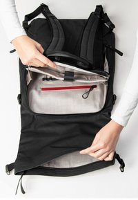 Zaino nero con spallacci imbottiti, un compartimento principale con zip che presenta un interno grigio e una tasca con zip rossa, mostrato con mani che ne dimostrano l'uso.