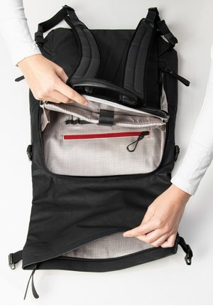 Schwarzer Rucksack mit gepolsterten Trägern, einem Hauptfach mit Reißverschluss, das ein graues Innenfutter und eine rote Reißverschlusstasche aufweist, präsentiert mit Händen, die die Verwendung demonstrieren.