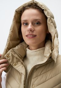 Beige, gepolsterte Weste mit hohem Kragen und Kapuze. Verfügbar mit goldfarbenen Hardwareelementen und gestepptem Textil. Getragen über einem cremefarbenen, gerippten Pullover.