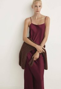 Vestido de satén burdeos con tirantes finos, combinado con un cárdigan de punto texturizado en marrón. La tela cae suavemente, creando una silueta elegante.