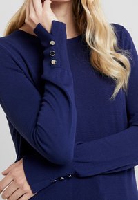 Top de manga larga azul marino en tejido de punto con relieve. Presenta un cuello redondo y tres botones decorativos en cada puño.