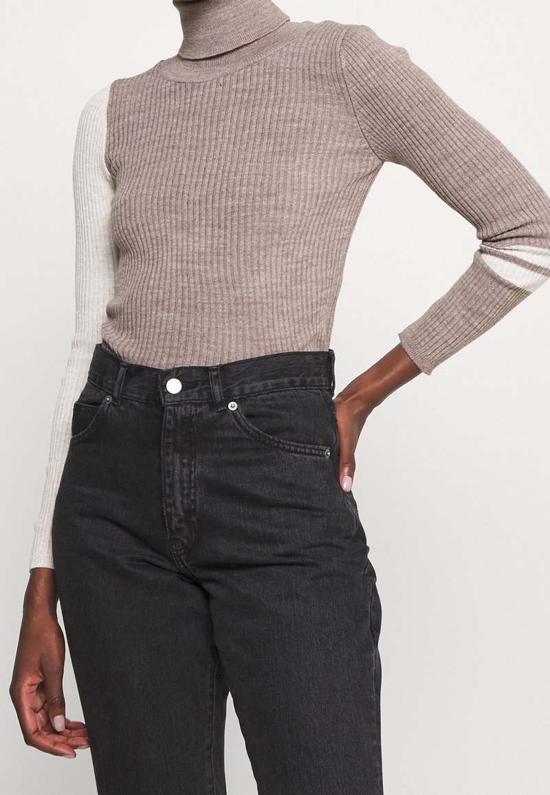 Pull à col roulé côtelé à manches longues, beige et crème, associé à un jean noir taille haute, porté par une personne avec la main sur la hanche.