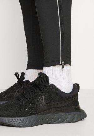 Schwarze Sportschuhe mit strukturiertem Mesh-Obermaterial, Gummisohle und Schnürsenkeln. Kombiniert mit weißen gerippten Socken und schwarzen schmal geschnittenen Hosen mit Reißverschlussdetails.