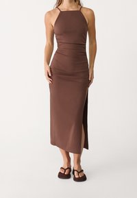 Brun, tætsiddende maxikjole med lige halsudskæring, tynde stropper og slids i siden. Blødt, tekstureret stof, båret med mørke sandaler.