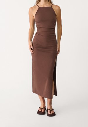 Bruine, aansluitende maxi-jurk met een rechte halslijn, dunne bandjes en een zijsplit. Zachte, gestructureerde stof, gedragen met donkere sandalen.