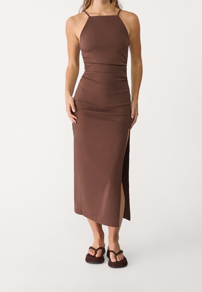 Brun, tætsiddende maxikjole med lige halsudskæring, tynde stropper og slids i siden. Blødt, tekstureret stof, båret med mørke sandaler.