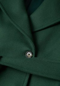 Manteau en laine vert foncé avec un bouton noir et argenté étiqueté "MAG MANG", montrant le col replié et la texture du tissu.