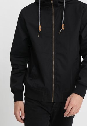 Zwarte hoodie met ritssluiting en trekkoorden, voorzien van bruine leren accenten, gedragen door een persoon met zichtbare handen langs de zij.