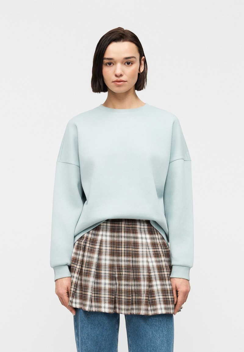Femme portant un sweat-shirt oversize bleu clair, mini-jupe plissée à carreaux par-dessus un jean bleu, debout devant un fond blanc.