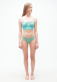 Γυναίκα που στέκεται ξυπόλητη, φοράει πράσινο και μπλε καρώ cropped τοπ και μονόχρωμα πράσινα μαγιό κάτω μέρη, κοιτάζοντας προς τα δεξιά της, με φόντο ένα απλό υπόβαθρο.