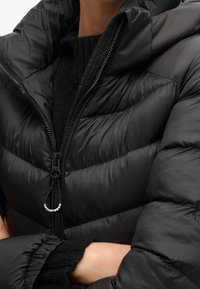 Persona che indossa una giacca a vento nera parzialmente zipata su un maglione nero a maglia, braccia incrociate, con il tirazip etichettato "Superdry" visibile.