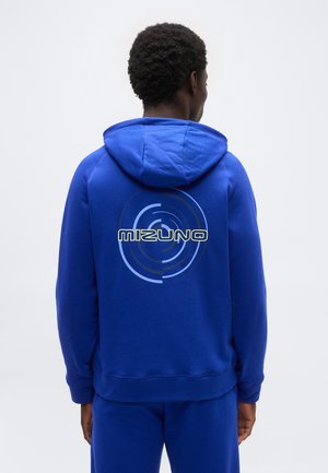 Man in een blauwe hoodie en broek, staand met de rug naar de kijker, met een cirkelvormig ontwerp en de tekst "Mizuno" op de achterkant van de hoodie.