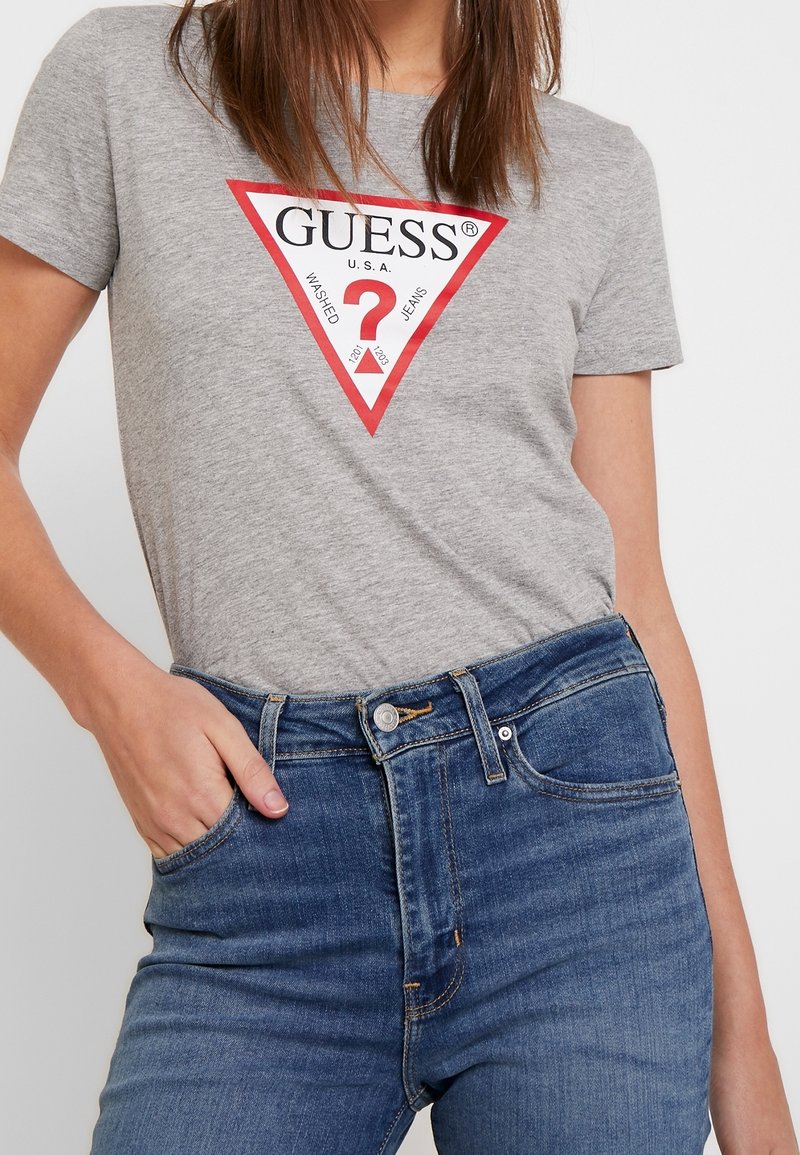 Graues Baumwoll-T-Shirt mit einem roten dreieckigen Logo, das "GUESS" und ein Fragezeichen zeigt. Kombiniert mit high-waisted blauen Denim-Jeans.