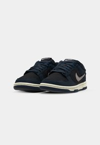 Námořnické modré tenisky s texturovanou tkaninou, s lehce šedým logem Nike, plochými tkaničkami a bílou mezipodešví s vzorovanou podešví.