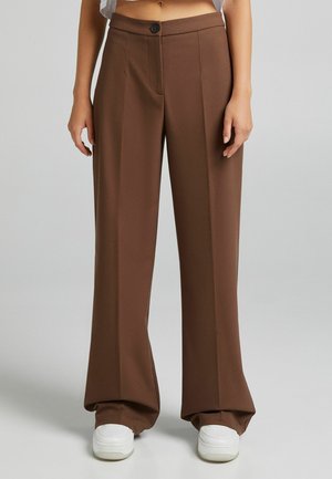 Pantalons larges marron au toucher lisse, dotés d'une fermeture à bouton à l'avant et de deux poches latérales. Associés à des baskets blanches.