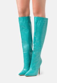 Botas turquesas hasta la rodilla, en piel brillante con estampado de cocodrilo en relieve, con punteras puntiagudas y altos tacones de aguja.