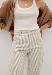 Pruhované béžové kalhoty s vysokým pasem, s knoflíkovým zapínáním a bočními kapsami. V kombinaci s bílým tílkem a hnědou košilí.