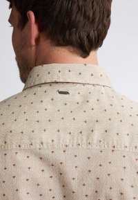 Lichtbeige shirt met een textuurpatroon, voorzien van kleine bruine bloemenaccenten en een label aan de achterkant van de kraag. Casual ontwerp, met kraag.
