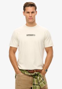 Camiseta corta de manga en color crema con un logo negro de "SUPERDRY". Combinada con pantalones caqui y una camisa de cuadros verde atada en la cintura.