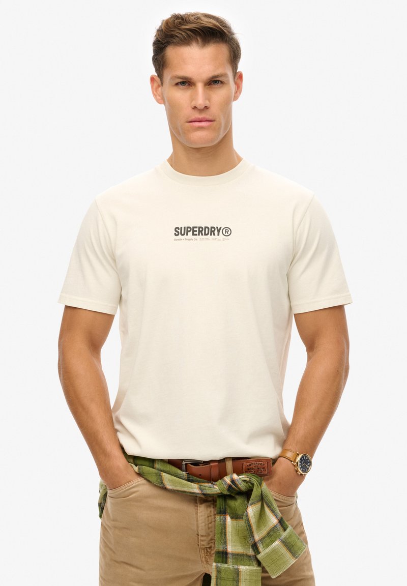 Cremefarvet T-shirt med korte ærmer og et sort "SUPERDRY" logo. Båret med khaki bukser og en grønternet skjorte, bundet i taljen.