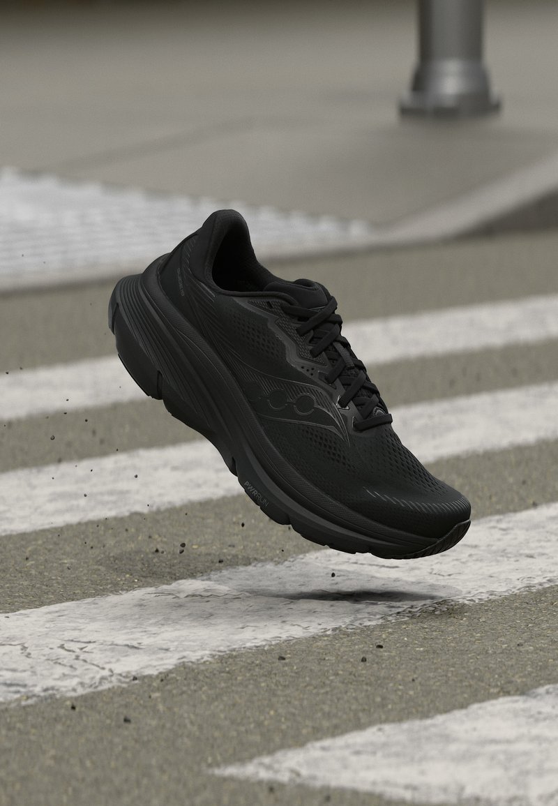 Μαύρο αθλητικό παπούτσι που τρέχει στον αέρα πάνω από διάβαση πεζών σε ασφαλτοστρωμένο δρόμο, με μικρά υπολείμματα να πετάγονται από κάτω του.