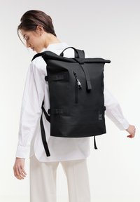 Czarny plecak typu roll-top wykonany z trwałego materiału, z regulowanymi paskami, przednią kieszenią na zamek i logo "GOT BAG" po bokach.