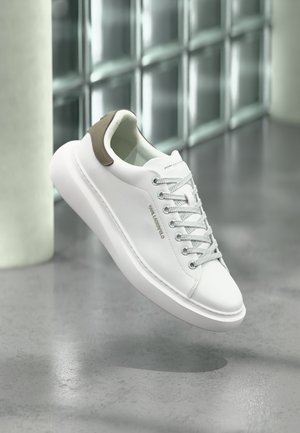 Λευκό δερμάτινο αθλητικό παπούτσι με χοντρή σόλα και γκρίζα κορδόνια, μπεζ λουράκι στη φτέρνα με την ετικέτα «KARL LAGERFELD», αιωρούμενο πάνω από γκρι πάτωμα με φόντο τοίχο από γυάλινους κύβους.