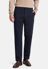 Marineblaue Baumwollhose mit flacher Vorderseite, Seitentaschen und lockerer Passform. Kombiniert mit braunen Leder-Loafers und einem beigen Pullover.