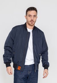 Marine bomberjack met geribbelde manchetten en tailleband, voorzien van een ritszak op de mouw en een oranje label op de voorkant.