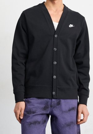 Mężczyzna w czarnym, zapinanym na guziki kardiganie Nike z białym logo na białej koszuli i fioletowych spodniach tie-dye, stoi z luźno opartymi rękami.