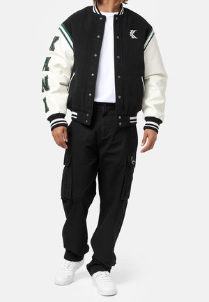 Giacca varsity bianca e nera con strisce verdi, bottoni a pressione e lettering sulle maniche. Abbinata a pantaloni cargo neri e sneakers bianche.