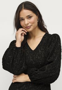 Abito nero con motivo floreale testurizzato, scollatura a V profonda e maniche a sbuffo. Tessuto liscio con una vestibilità strutturata. Modella in posa con la mano sul mento.