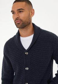 Cárdigan de punto en color marino con cuello de chal, patrón texturizado y cinco botones. Lleva puesto sobre una camiseta de cuello redondo blanca.