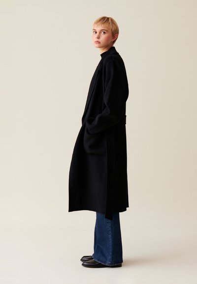 Cappotto nero realizzato in morbido tessuto, con una silhouette lunga, tasche laterali e un accento in vita grazie alla cintura. Abbinato a jeans a zampa d'elefante in denim scuro e mocassini.