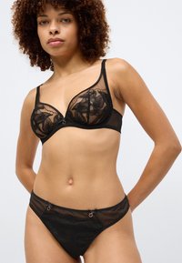 Czarny komplet bielizny koronkowej z przezroczystymi miseczkami z kwiatowym wzorem, regulowanymi ramiączkami oraz dekoracyjnymi elementami na majtkach.