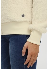 Kreemikasvalt fleece-jakk, millel on soonikkoes varrukad. Omab tekstuurset, karvast pinda ning väikest logot aktsendina hemil.