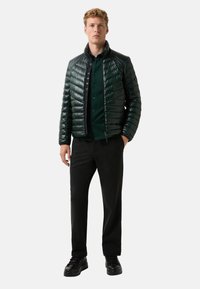 Junger Mann steht da und trägt eine dunkelgrüne, glänzende Steppjacke, ein grünes Hemd mit Knöpfen, schwarze Hosen und schwarze Schnürschuhe auf einem einfarbigen Hintergrund.