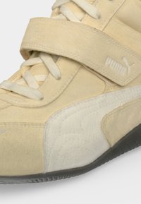 Gros plan d'une basket Puma beige montrant les lacets, une bride velcro avec le logo Puma et un panneau latéral blanc texturé sur une semelle noire.