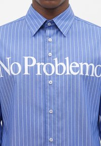 Chemise bleue à fines rayures avec boutons et texte blanc « No Problemo » sur la poitrine, portée par une personne.