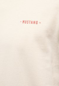 Tissu beige avec un petit texte rouge « 19 MUSTANG 32 » centré près du haut.