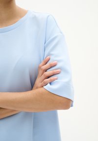Světle modrý top s krátkým rukávem, kulatým výstřihem a ohrnutými rukávy. Hladká textura, volný střih, který ukazuje ruku překříženou přes hruď.