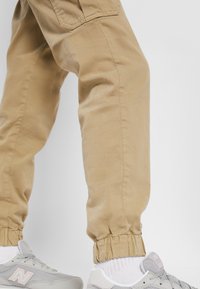 Khaki cargobyxor med elastiska muddar, tillverkade av ett tjockt bomullstyg. Har uttalade sidofickor. Kombineras med grå sneakers.