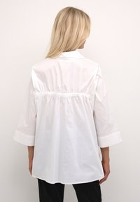 Camicia bianca in cotone con colletto, maniche a tre quarti e dettaglio della vita arricciato. Tessuto morbido e vestibilità rilassata.